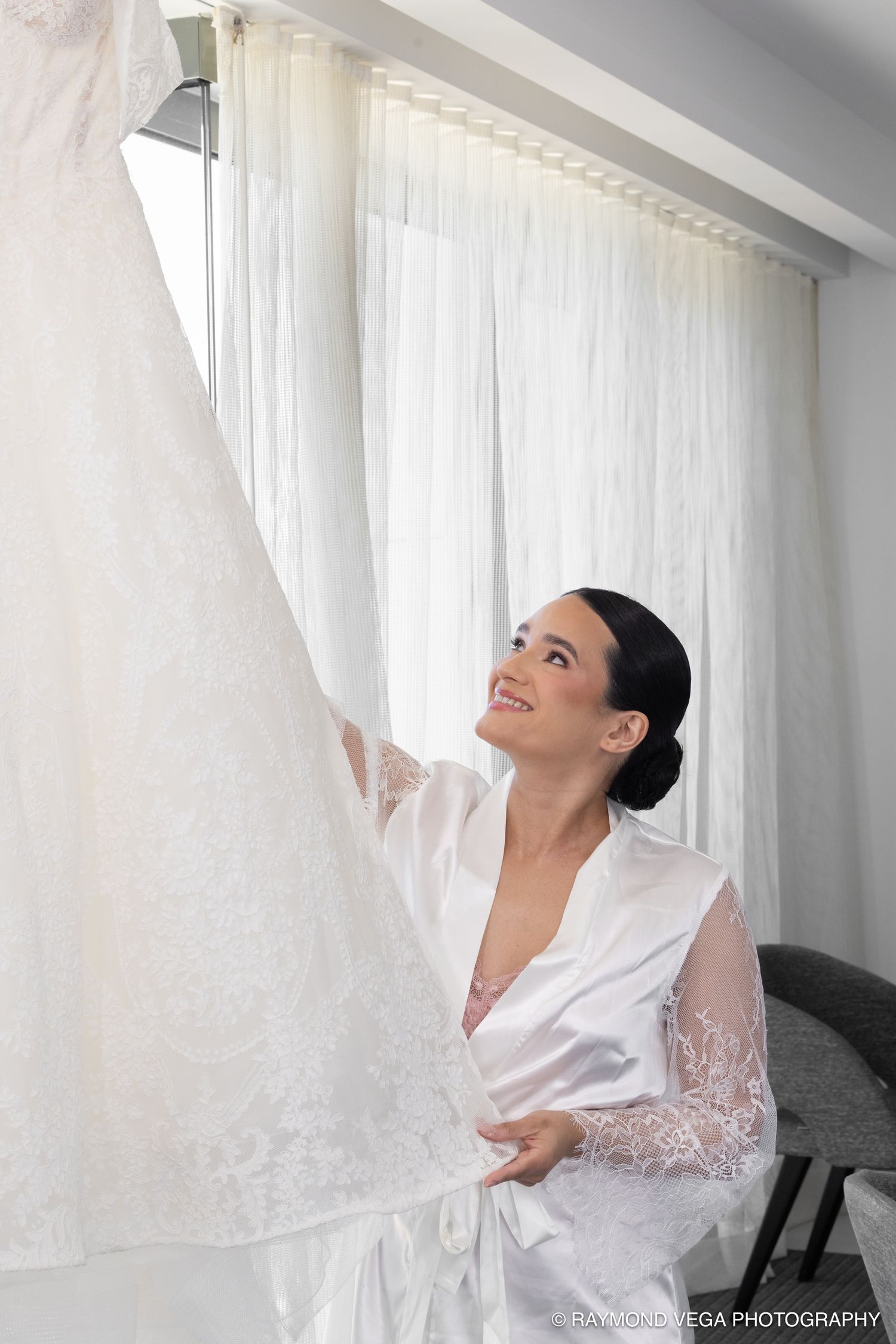 Smiling bride in white satin robe admiring her wedding dress hanging by a bright window.