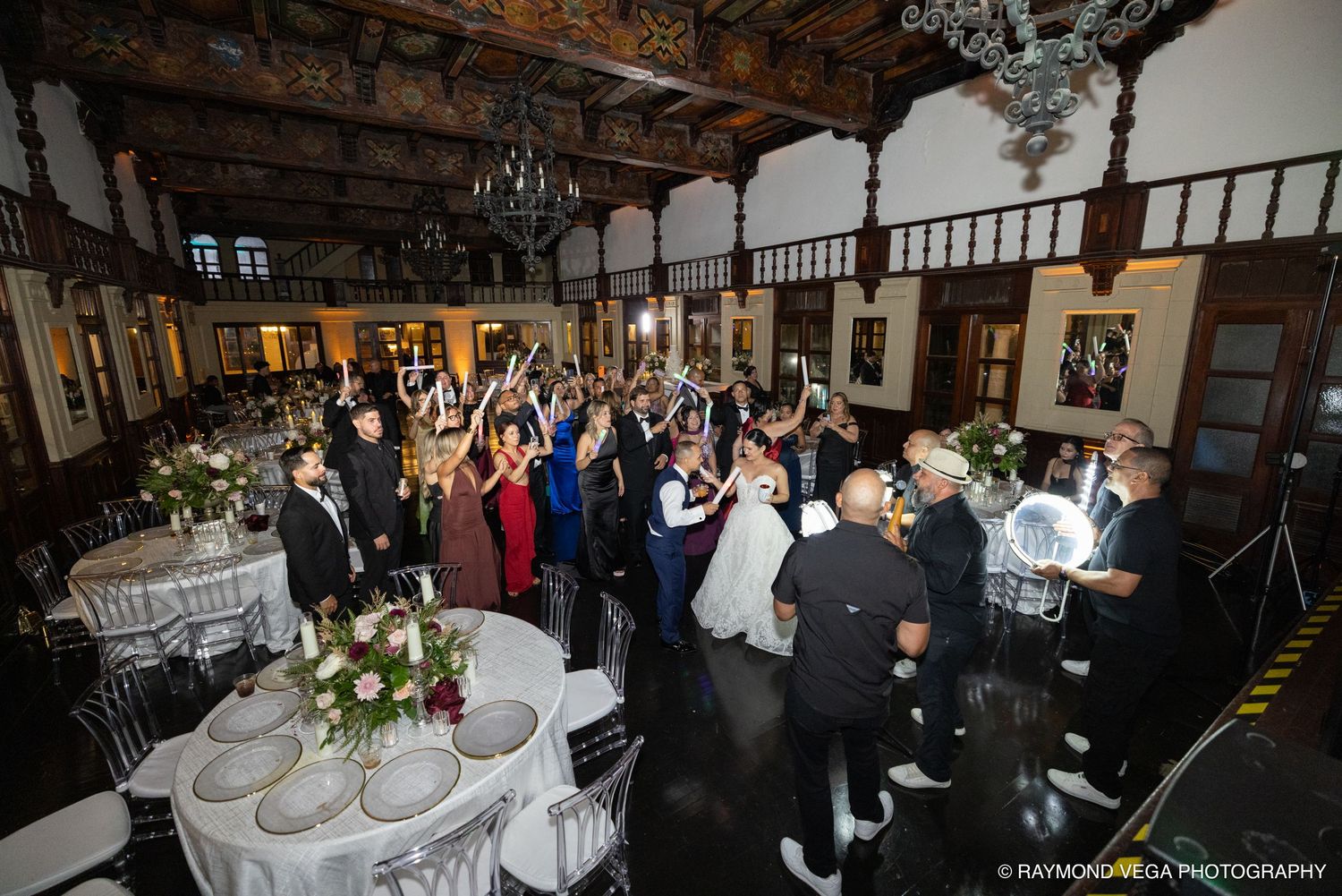 Elegant wedding reception with guests celebrating on the dance floor in a grand ballroom with chandeliers and floral centerpieces.