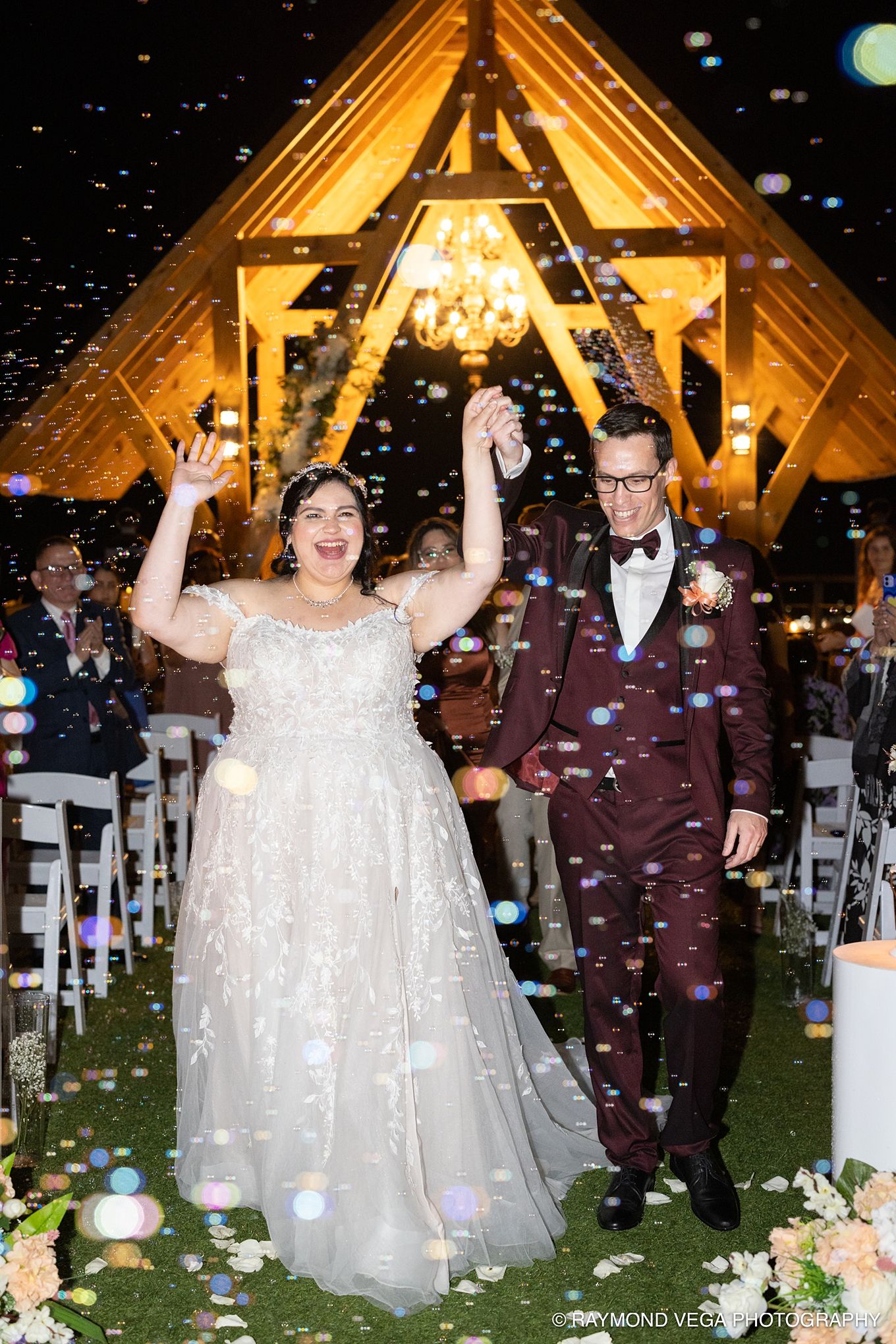 Joyful newlyweds celebrate recessional with bubbles at outdoor evening wedding ceremony with glowing A-frame chapel backdrop.