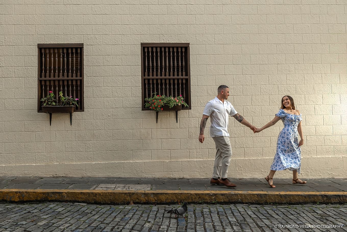Couple holding hands walking along a cobblestone street beside a white wall with barred windows and flower boxes.- Love Story