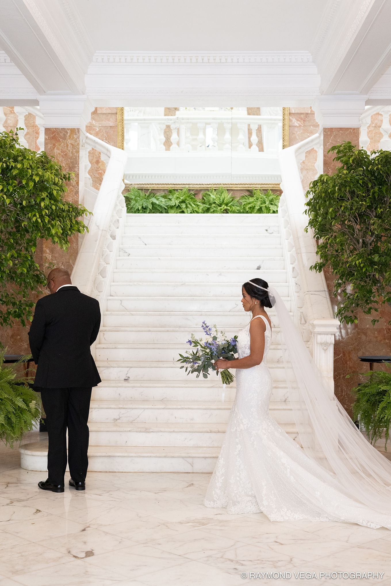 Bride in white lace gown with veil stands before marble staircase holding bouquet during elegant wedding first look.