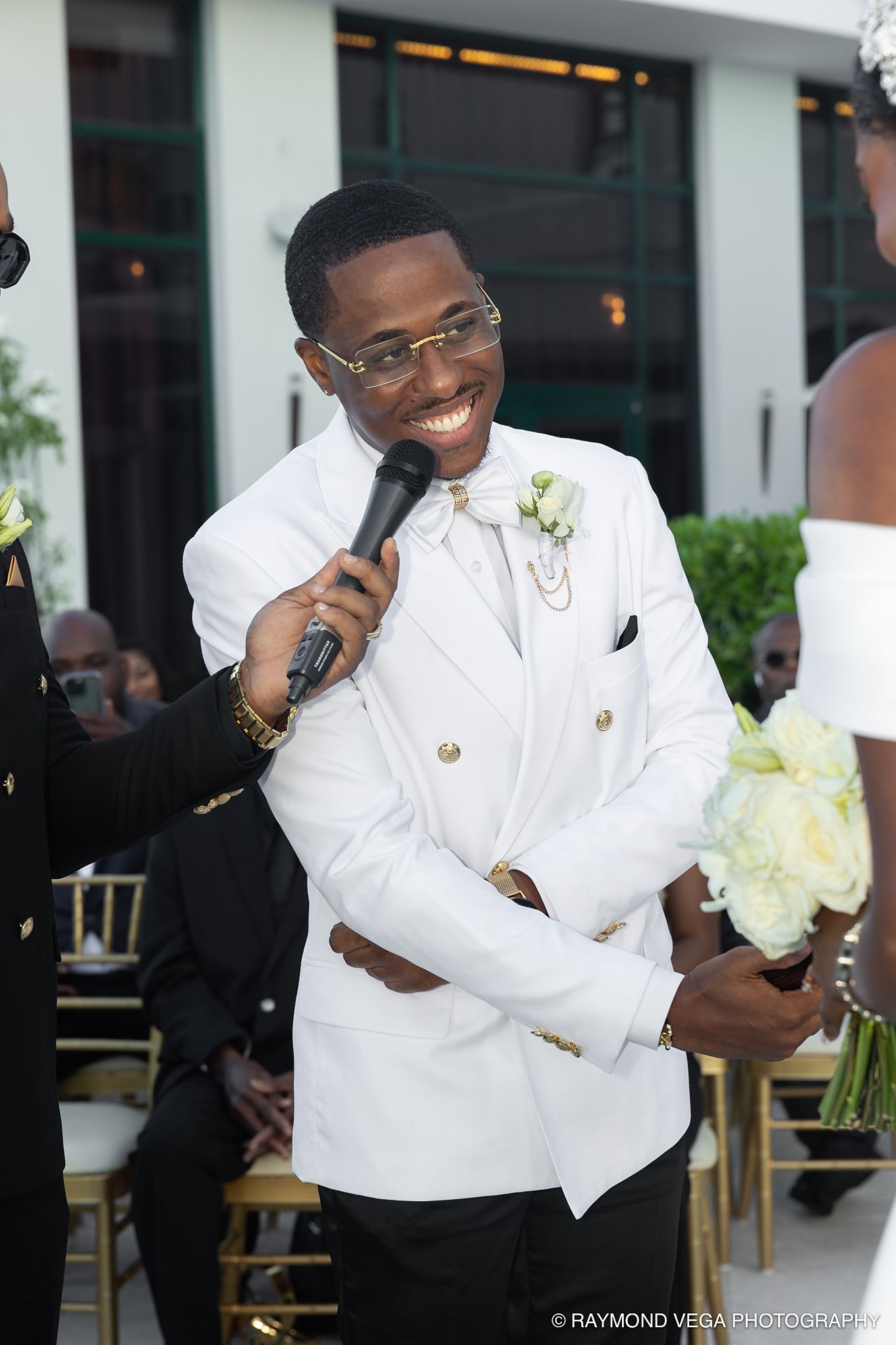 Smiling groom in white double-breasted suit holding microphone at outdoor wedding ceremony.