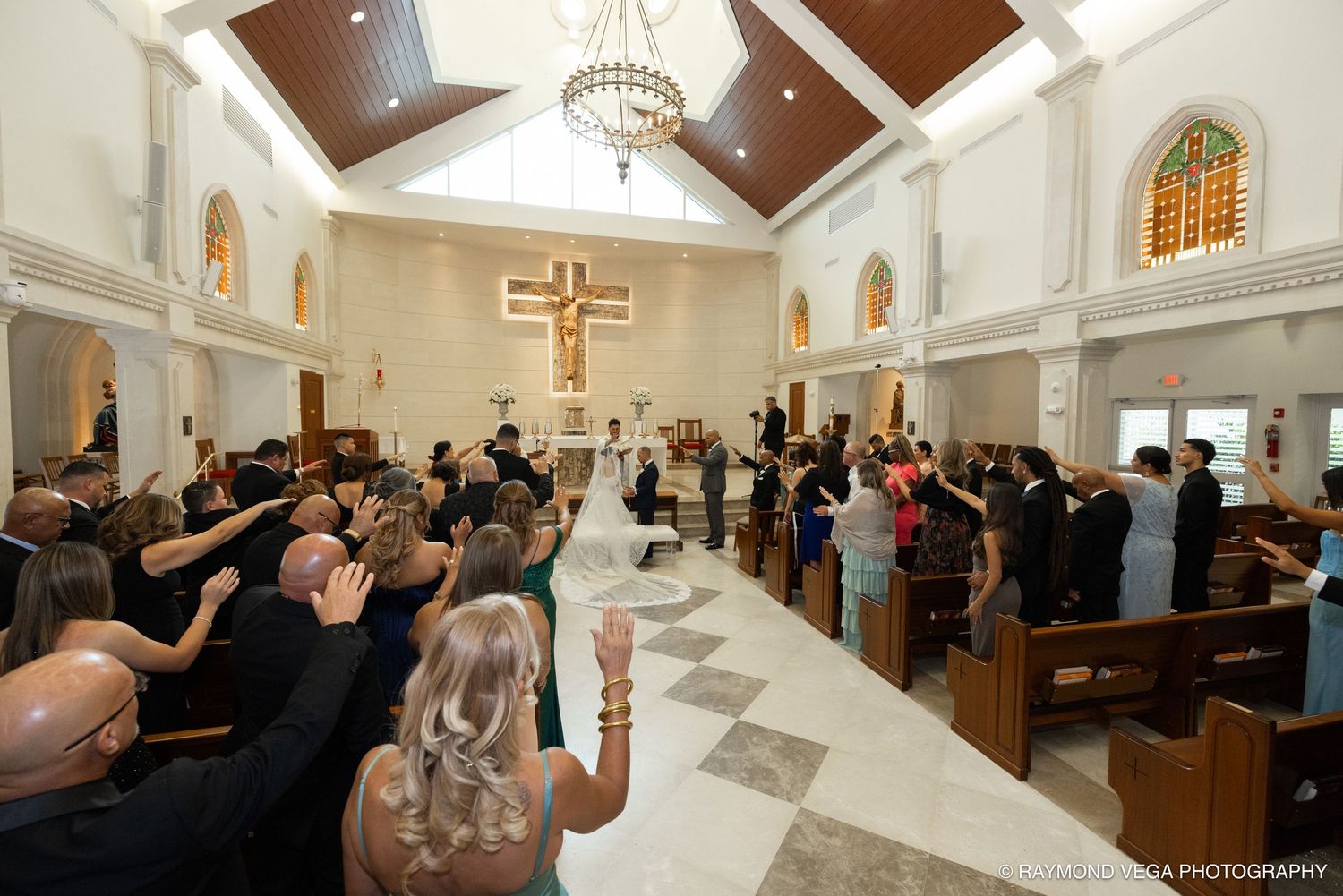 Wedding ceremony in a bright church with guests raising hands as couple stands at the altar beneath a wooden crucifix.