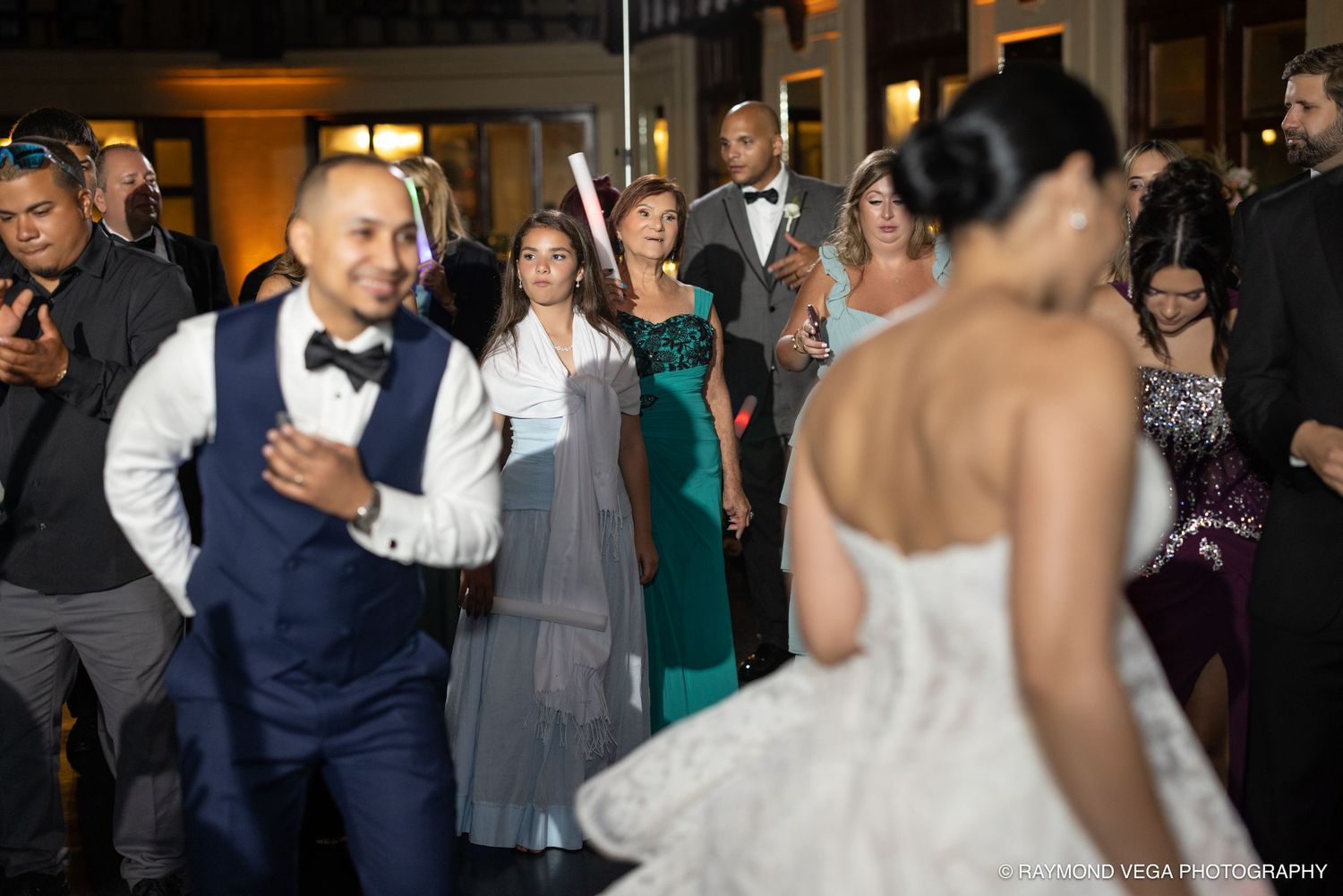 Groom in navy suit dances joyfully with bride in white wedding gown surrounded by guests at elegant reception.