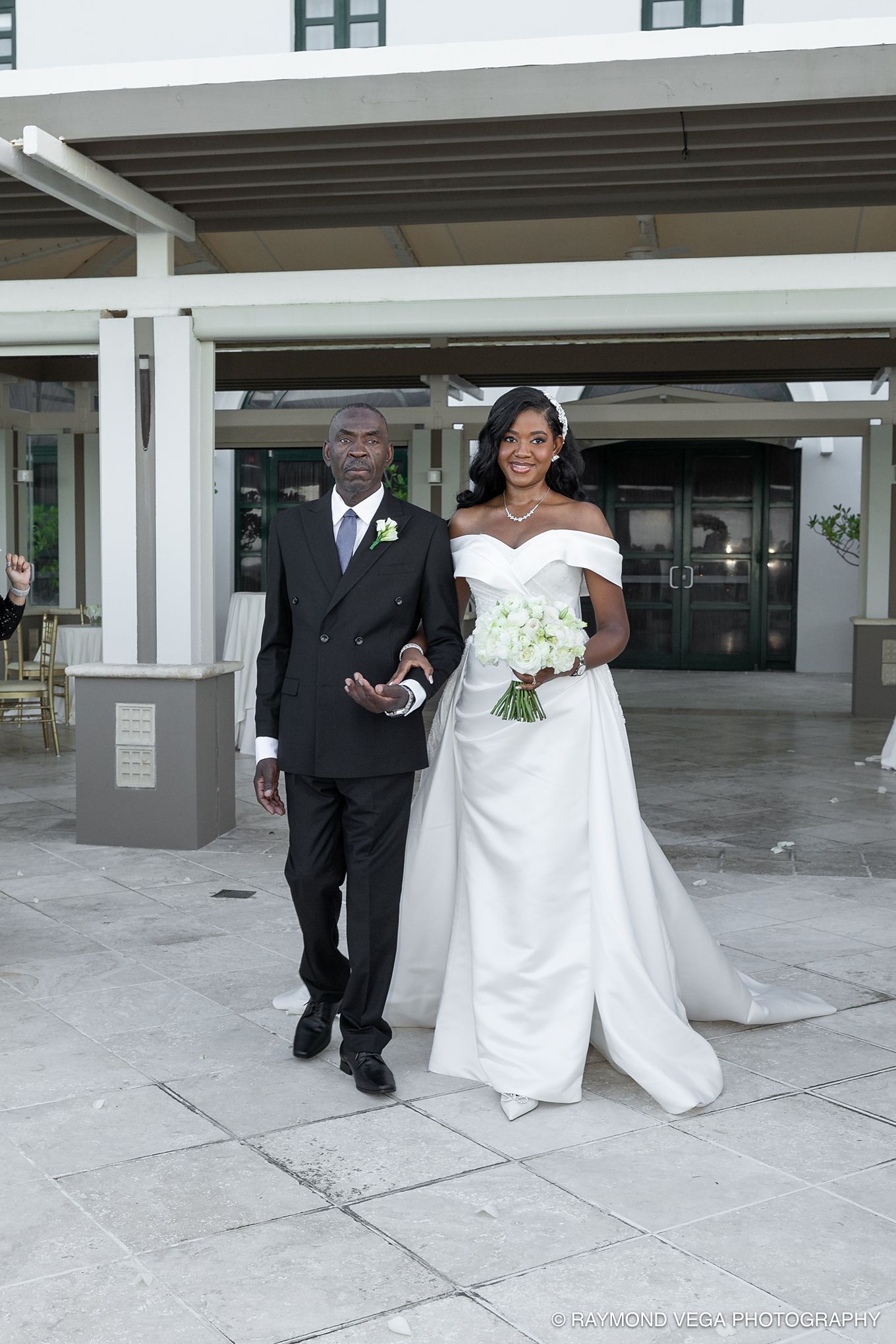 Bride in white off-shoulder gown holding bouquet walks with elder gentleman in black suit at outdoor wedding venue.