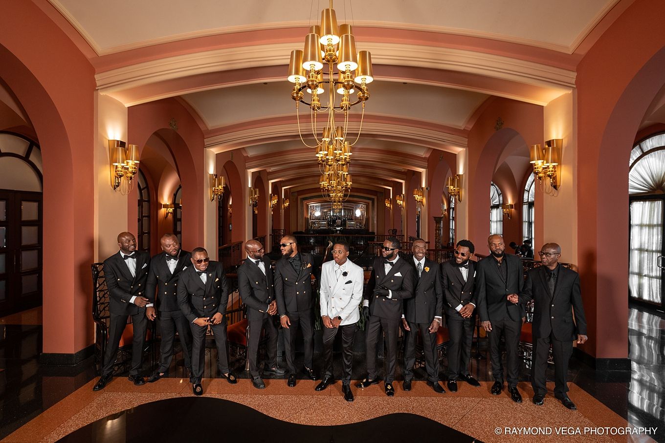 Groom in white jacket poses with groomsmen in black tuxedos in elegant venue hallway with chandelier. Hotel Condado Vanderbilt