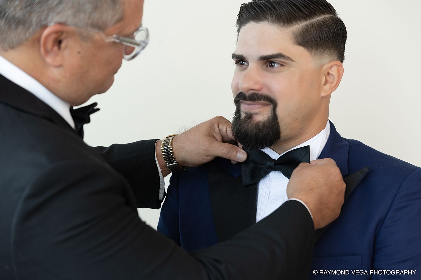 An older man adjusts the bow tie of a smiling groom in a navy tuxedo during wedding preparations.