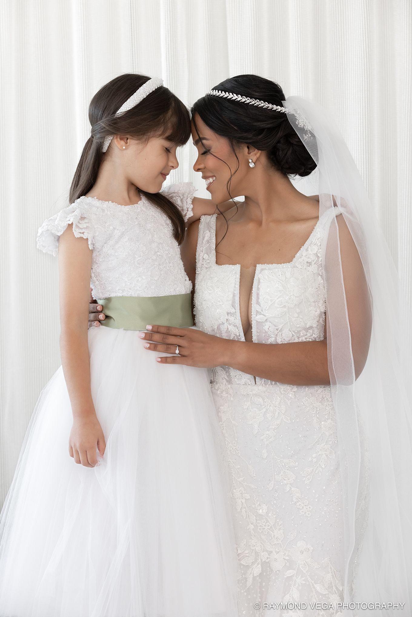 Bride in lace wedding gown and veil shares tender forehead-to-forehead moment with flower girl in white dress with sage sash.