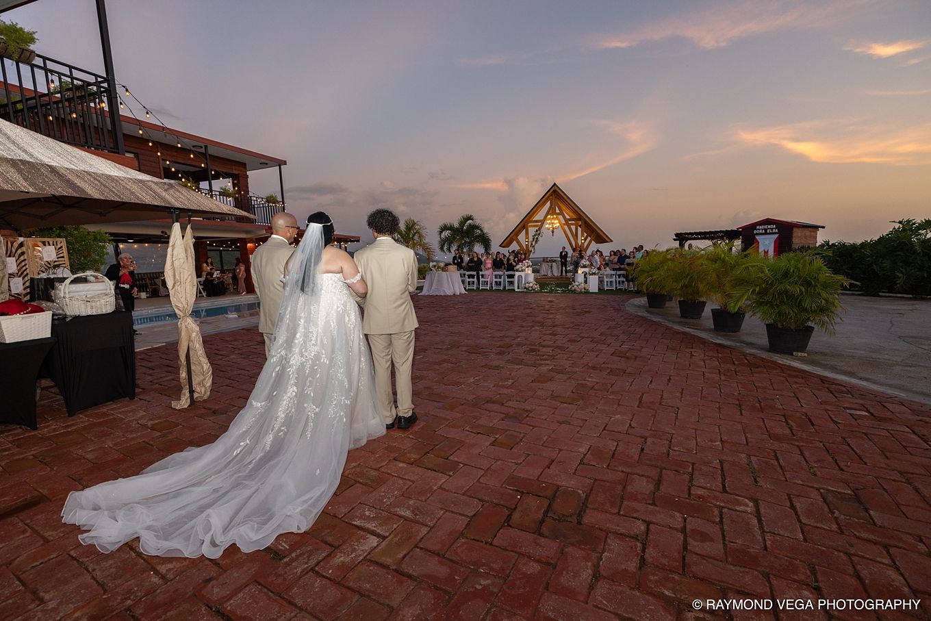 Bride and groom walking toward an outdoor wedding venue at sunset, with a beautiful brick courtyard and tropical scenery.