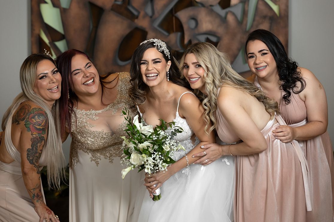 Bride with bouquet poses with four bridesmaids in blush and gold gowns, smiling together indoors.