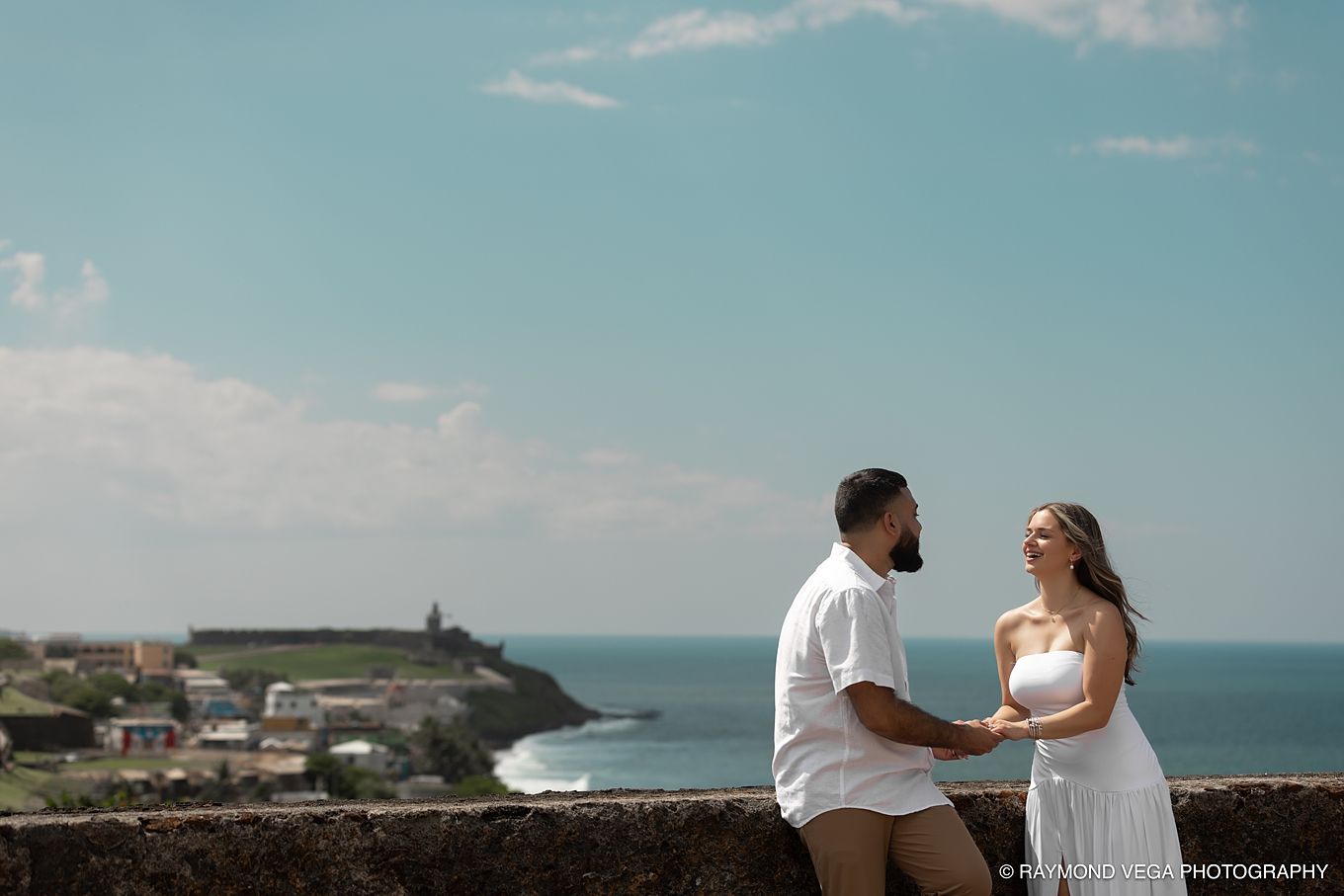 Couple holding hands on stone wall overlooking ocean and historic San Juan coastline under blue sky.