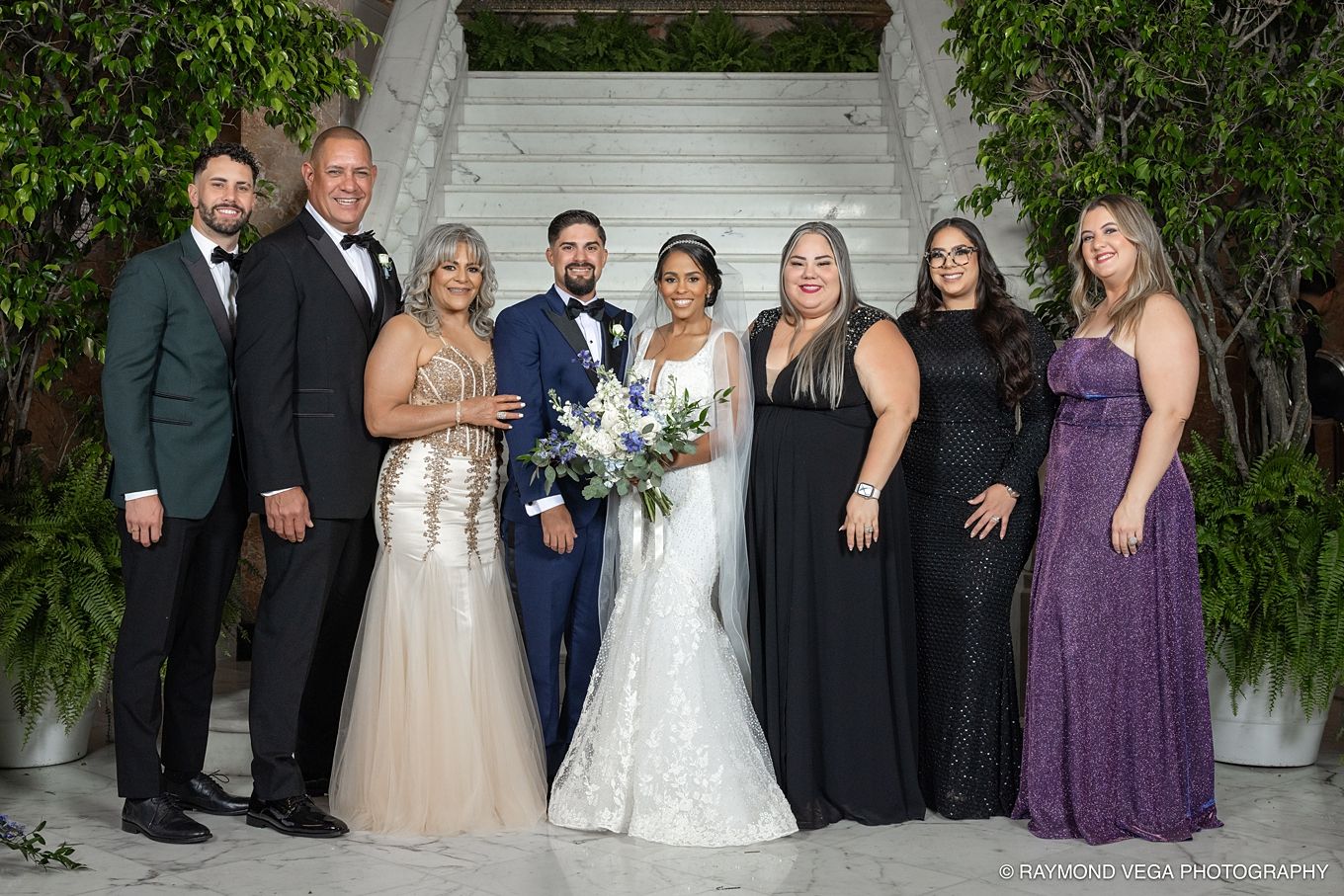 Wedding party poses on elegant staircase, bride in white gown with bouquet, groom in navy suit, family in formal attire. Antiguo Casino