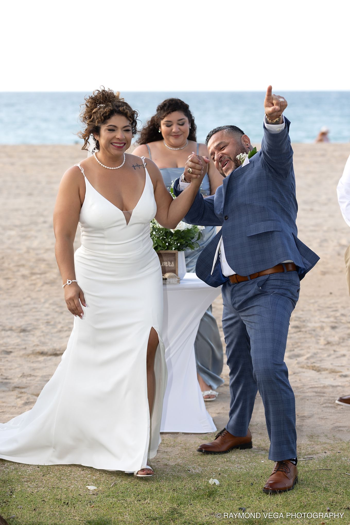 Joyful couple celebrates beach wedding, bride in white gown, groom in blue suit pointing up, officiant behind them.