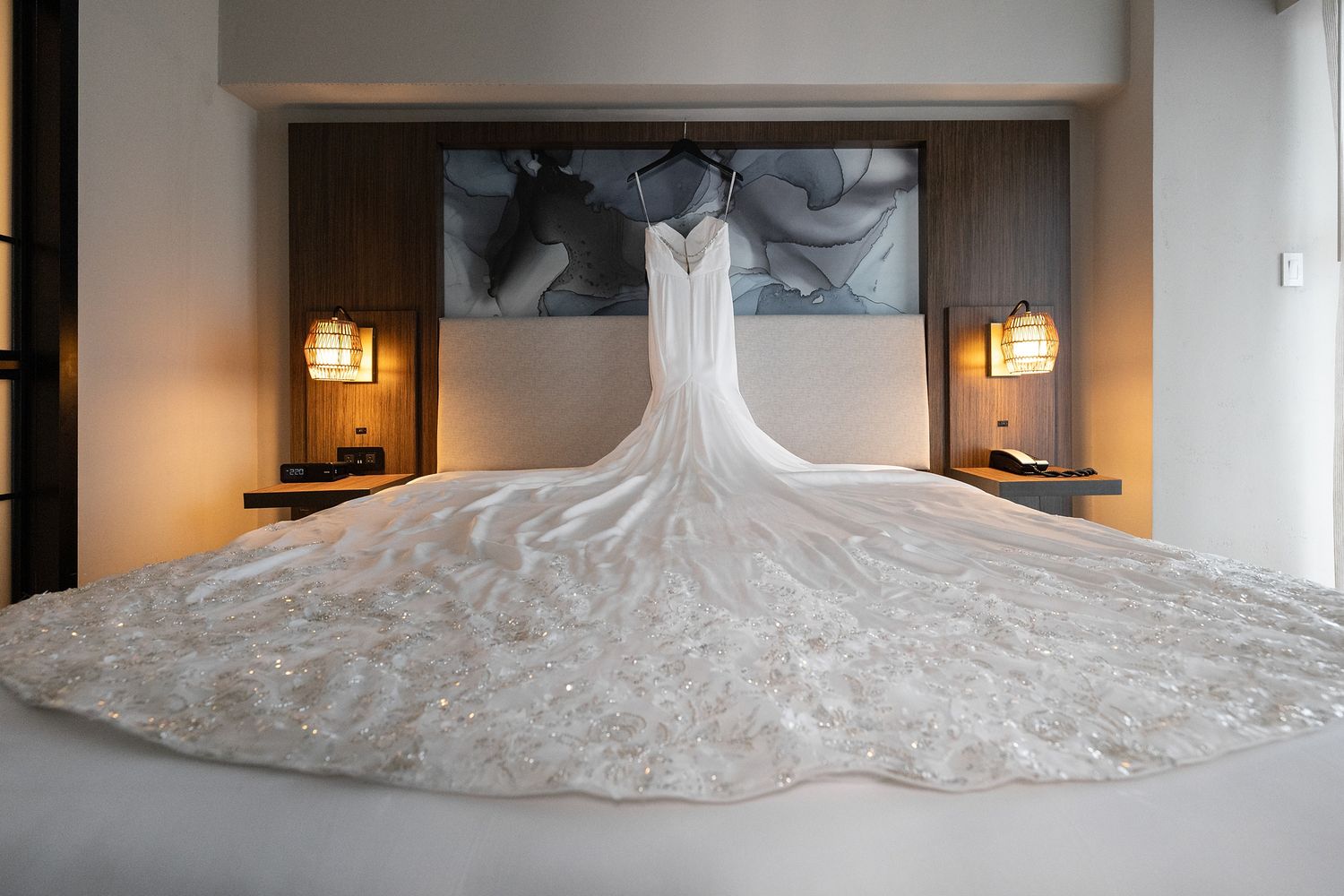 Elegant white lace wedding dress with a long cathedral train displayed on a hotel bed with warm ambient lighting.