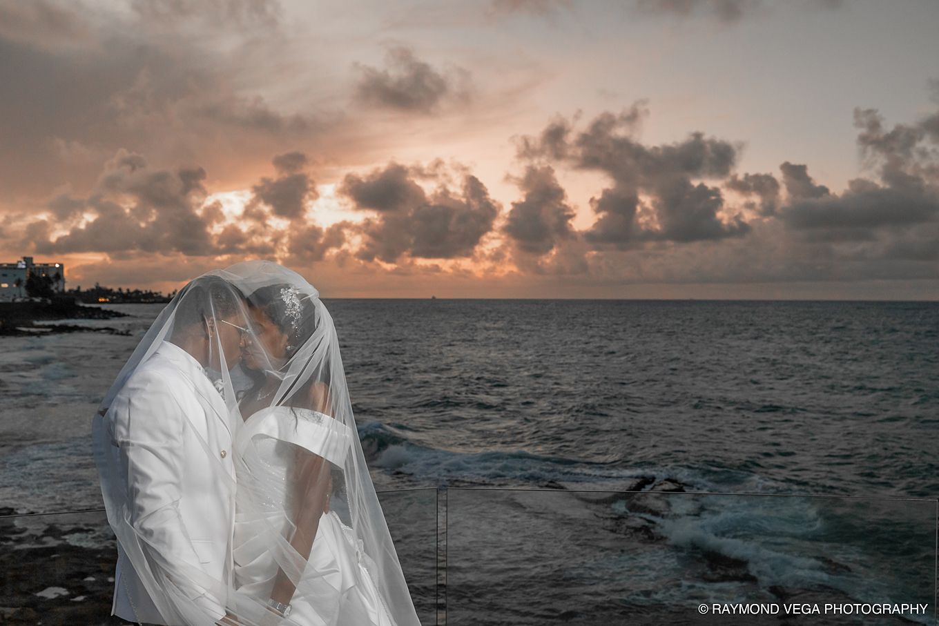 Bride and groom share romantic kiss under veil at ocean sunset, captured by Raymond Vega Photography.