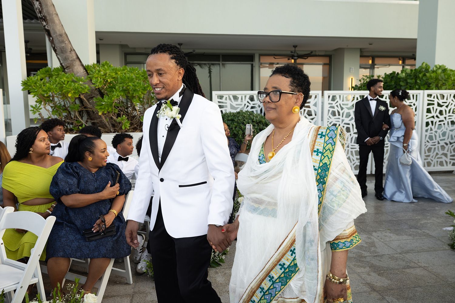 A groom in a white tuxedo walks hand-in-hand with a woman in a blue sari at an outdoor wedding ceremony.
