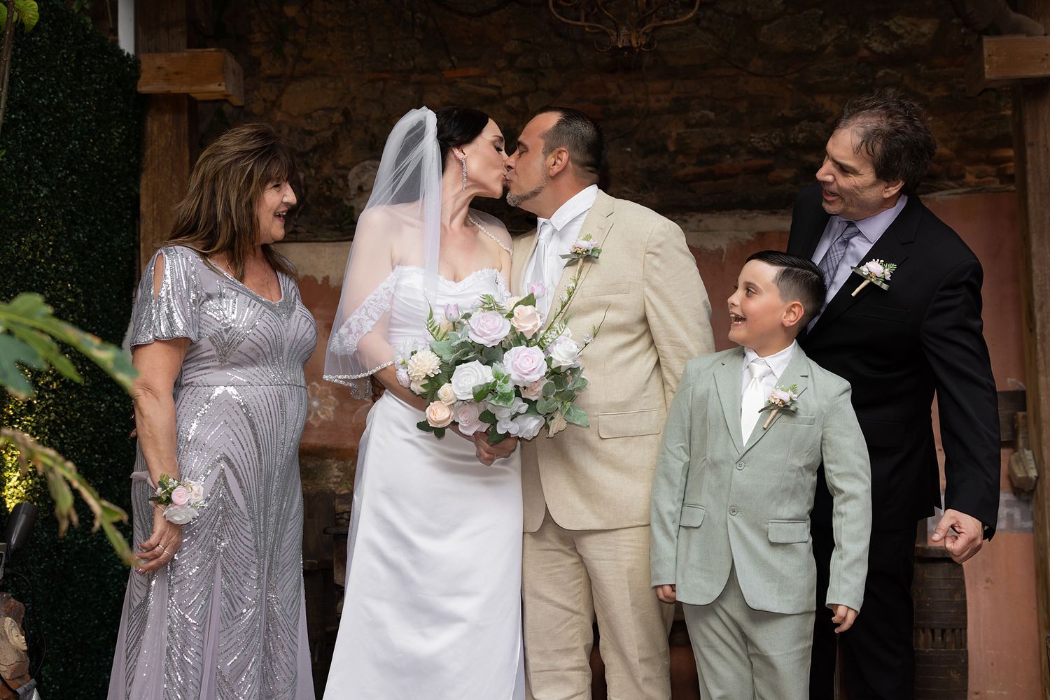 Bride and groom kissing at outdoor wedding ceremony with family including young boy in green suit and guests watching. at The Cannon Club