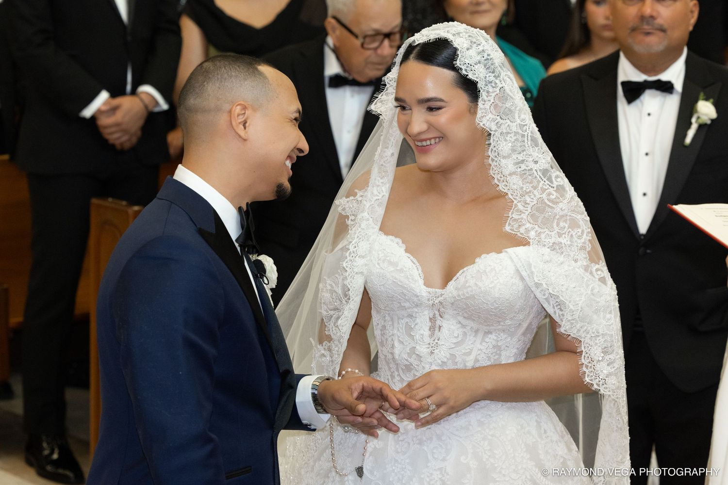 Bride in lace gown and veil smiles at groom in navy suit during elegant church wedding ceremony.