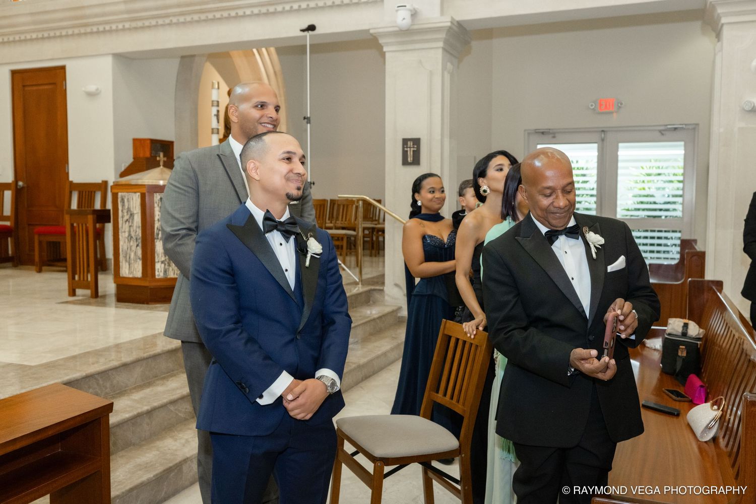 Groom in blue tuxedo smiles as guests gather in a church aisle during a wedding ceremony.
