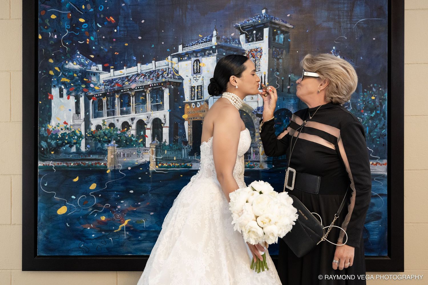 Bride in white gown holding bouquet stands with older woman in black near colorful painting at wedding venue.