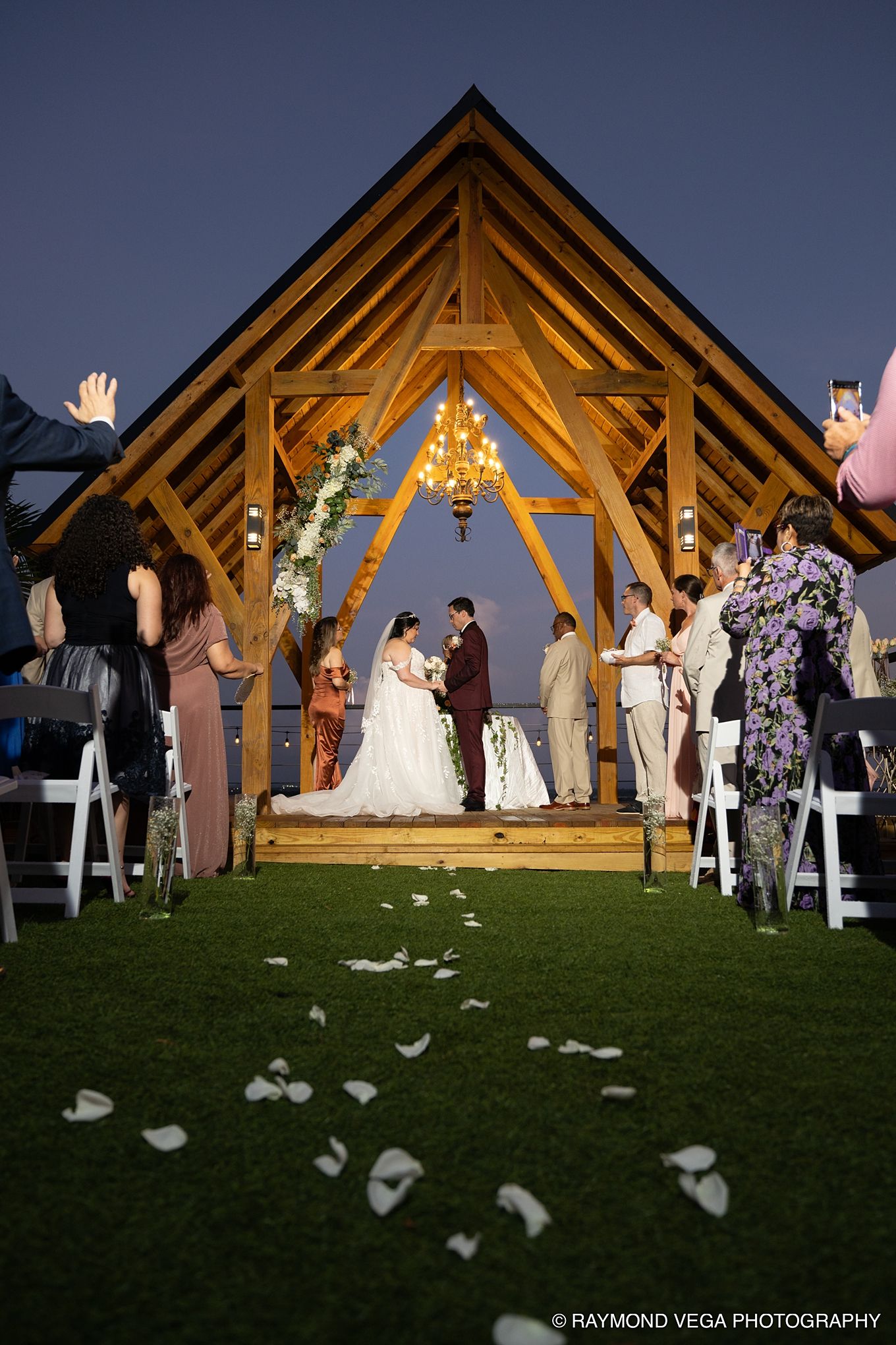 Outdoor wedding ceremony under a wooden pavilion with chandelier at dusk, white petals on grass aisle, guests standing.