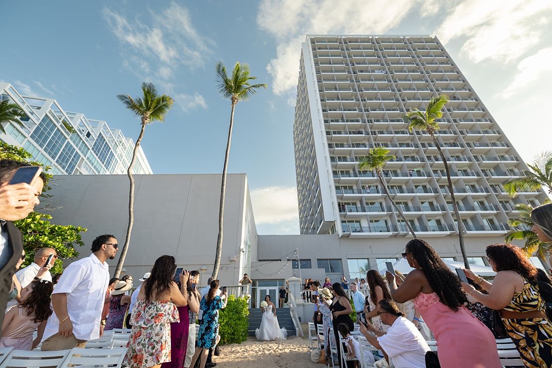 Outdoor wedding ceremony at a modern beachfront hotel with palm trees and guests seated under a clear blue sky. San Juan Marriott Resort