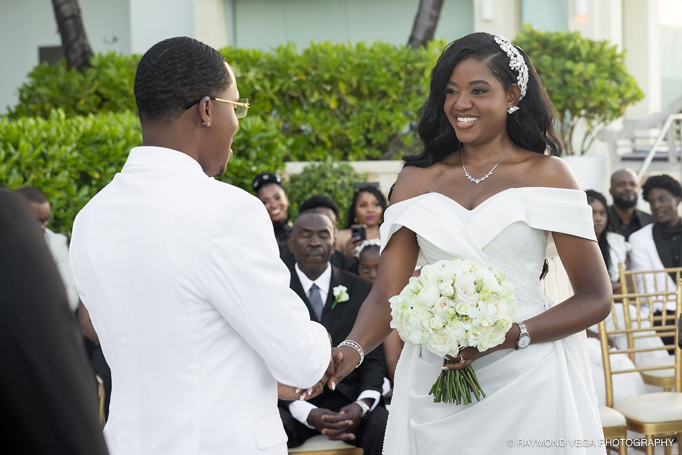 Bride and groom exchange vows at outdoor wedding ceremony, bride holding white bouquet in elegant off-shoulder gown.