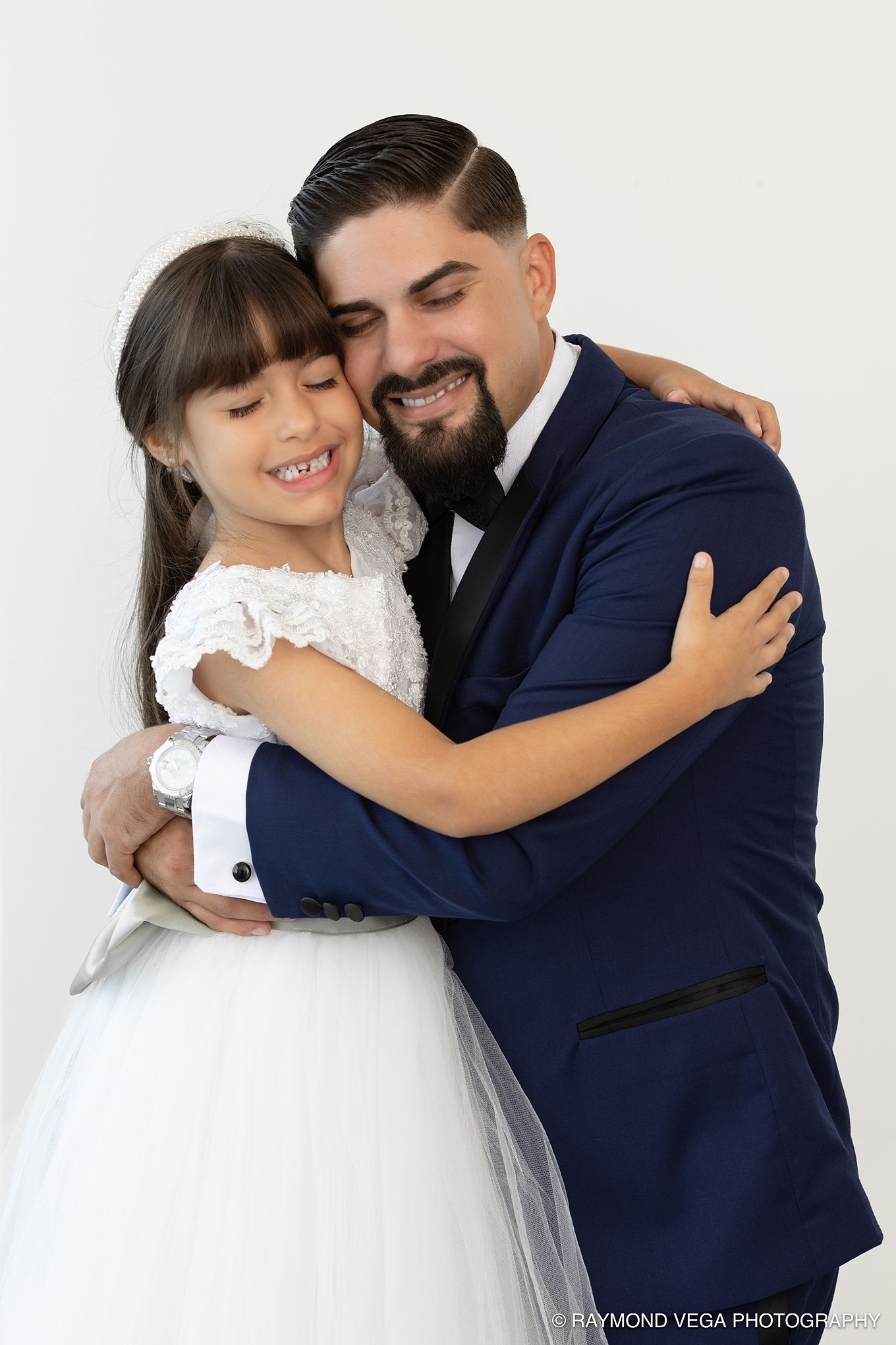 A man in a navy suit hugs a young girl in a white communion dress, both smiling warmly against a white background. Hotel la Concha Wedding