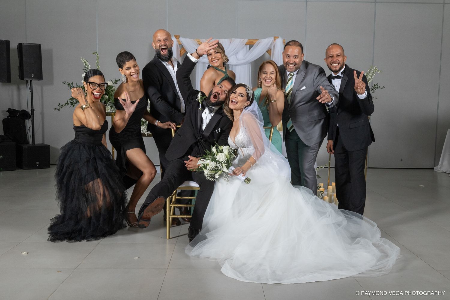 A joyful wedding party poses together, featuring a bride in a white gown and groom in a tuxedo surrounded by guests.