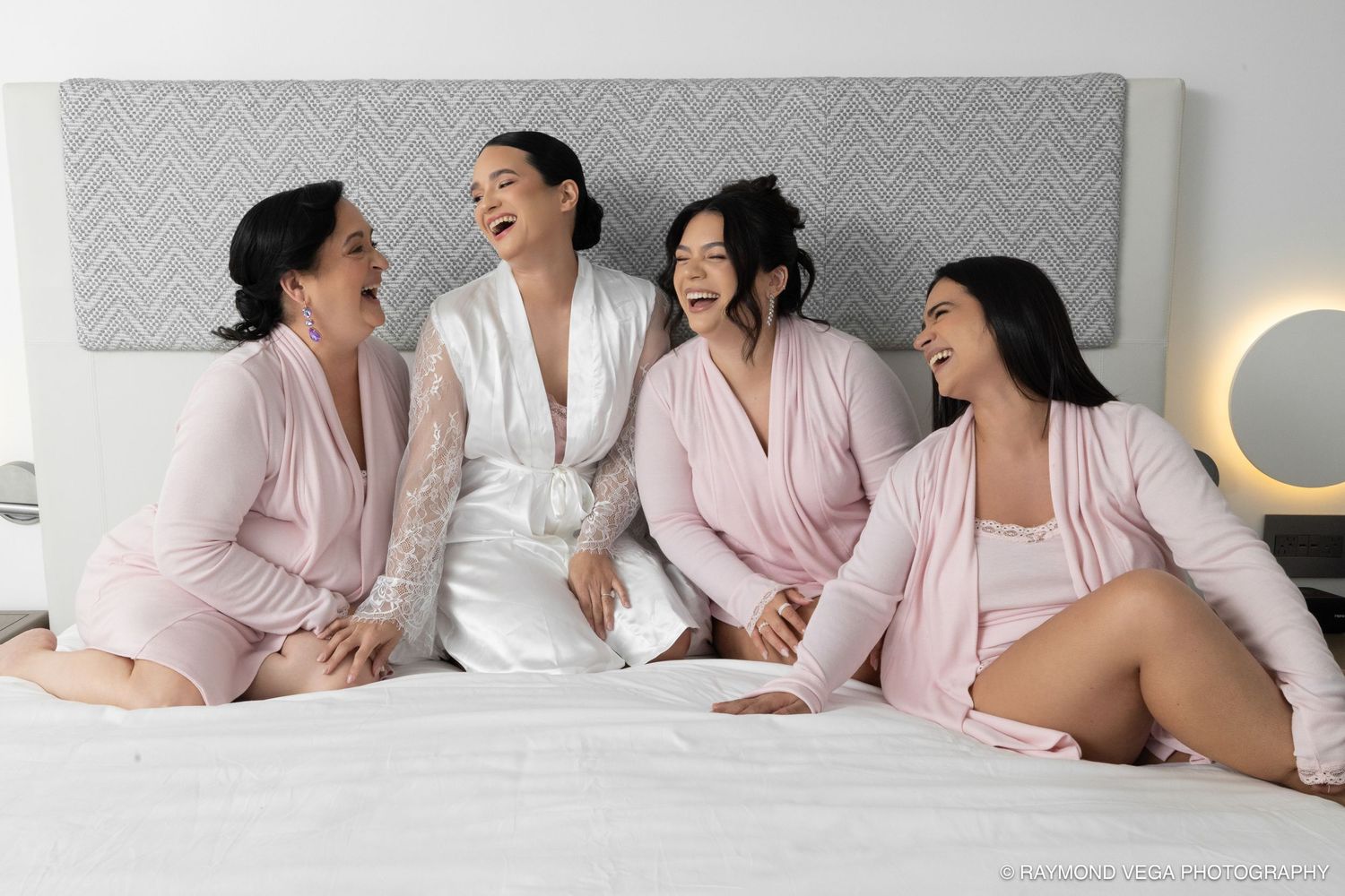 Four women in pink and white robes laughing together on a bed during bridal party getting ready.