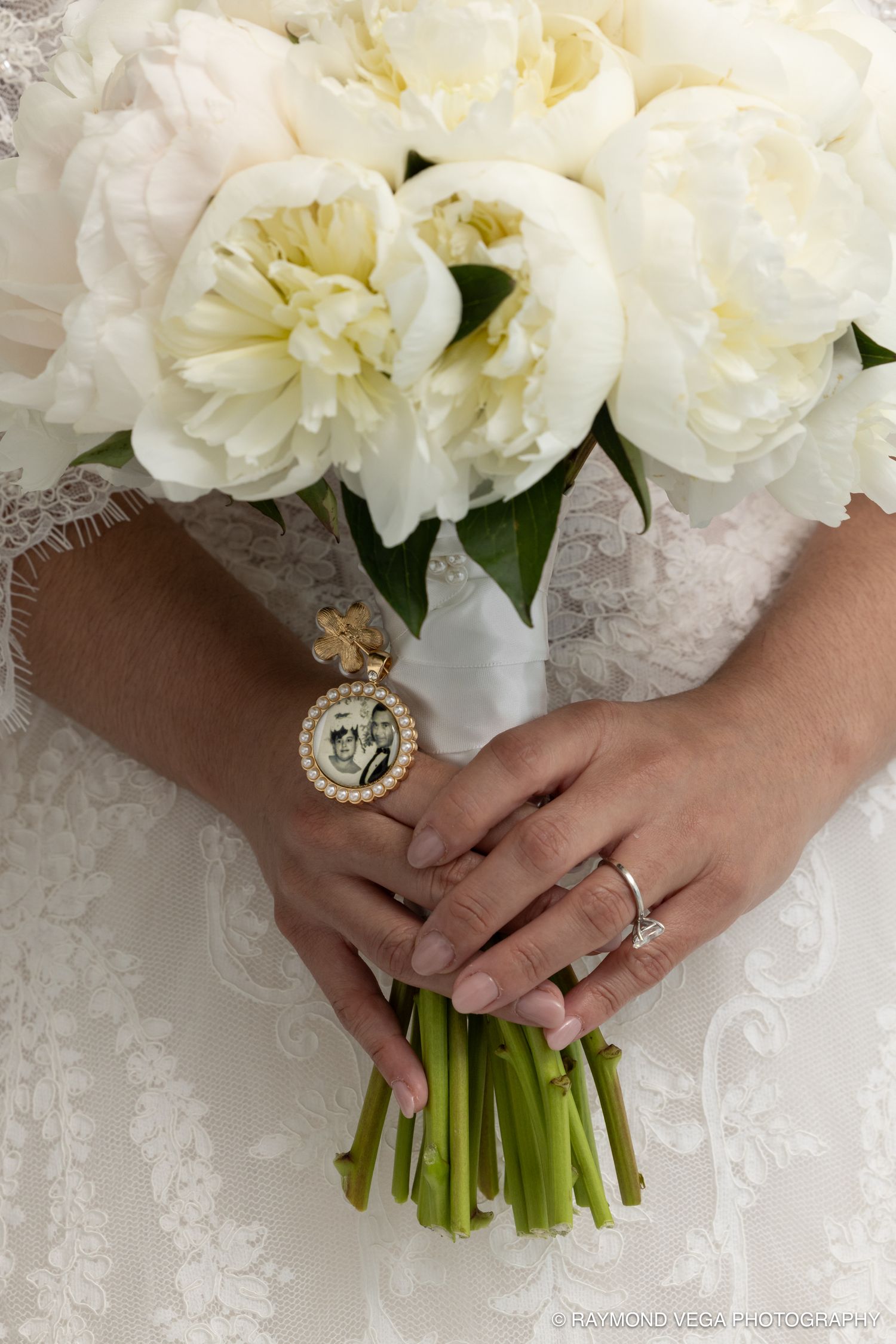 Bride holding white peony wedding bouquet with memorial photo charm, wearing diamond ring, lace dress detail visible.