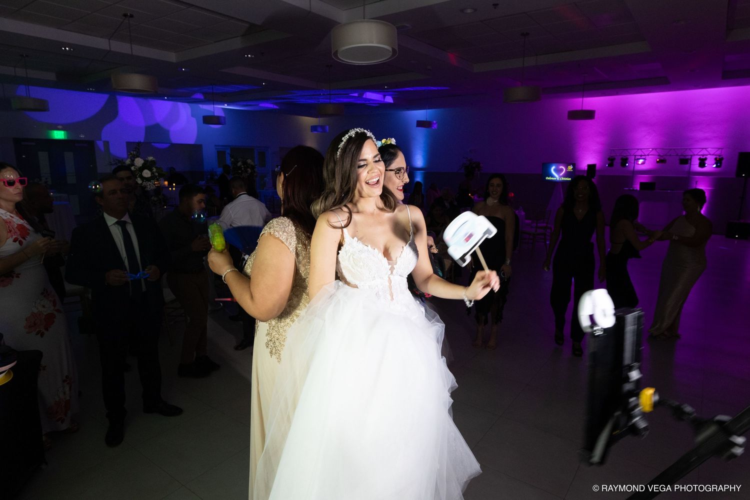 Bride in white wedding gown dancing joyfully with guests at a purple-lit wedding reception venue.