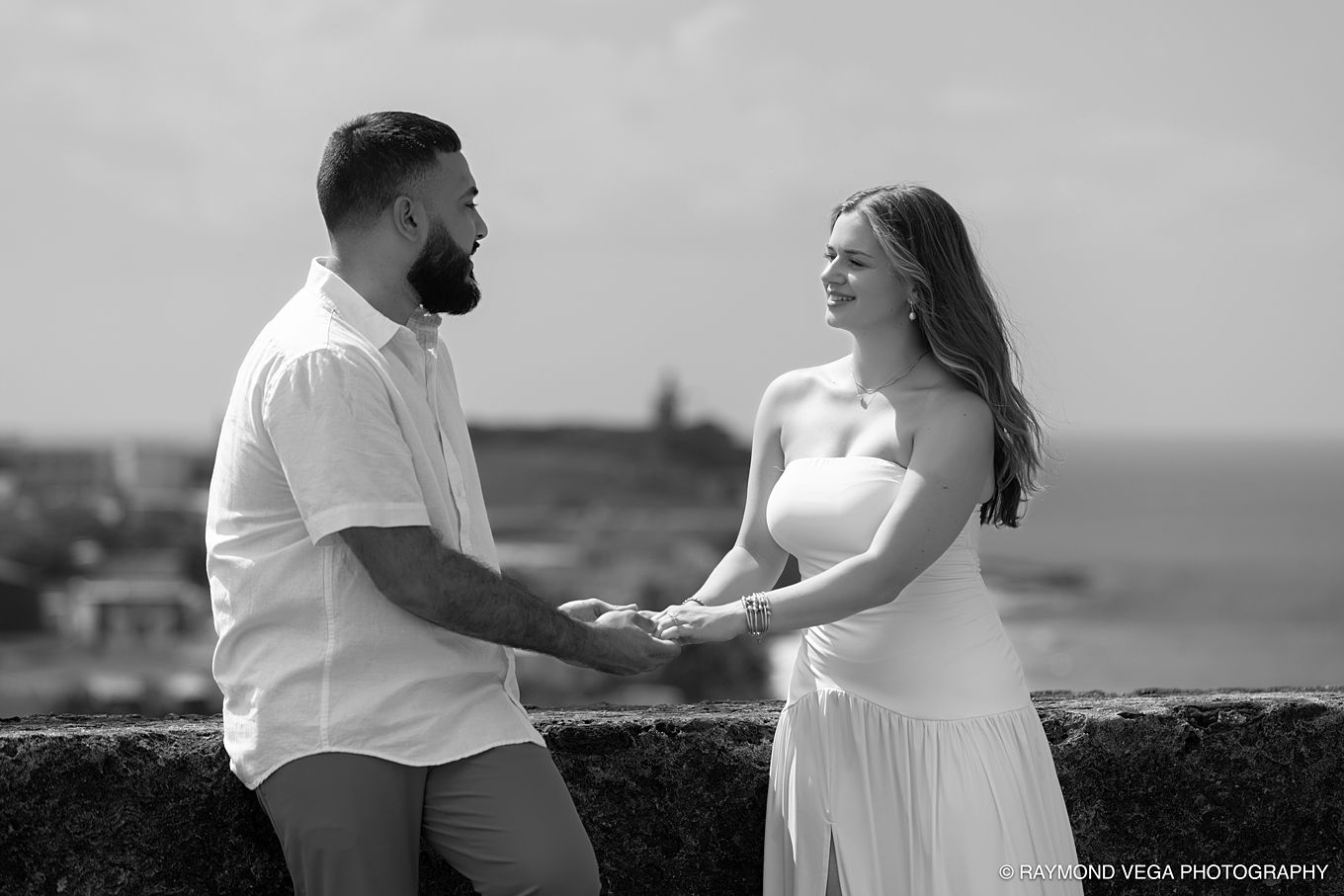 Black and white photo of a couple holding hands and smiling outdoors with a coastal town in the background.