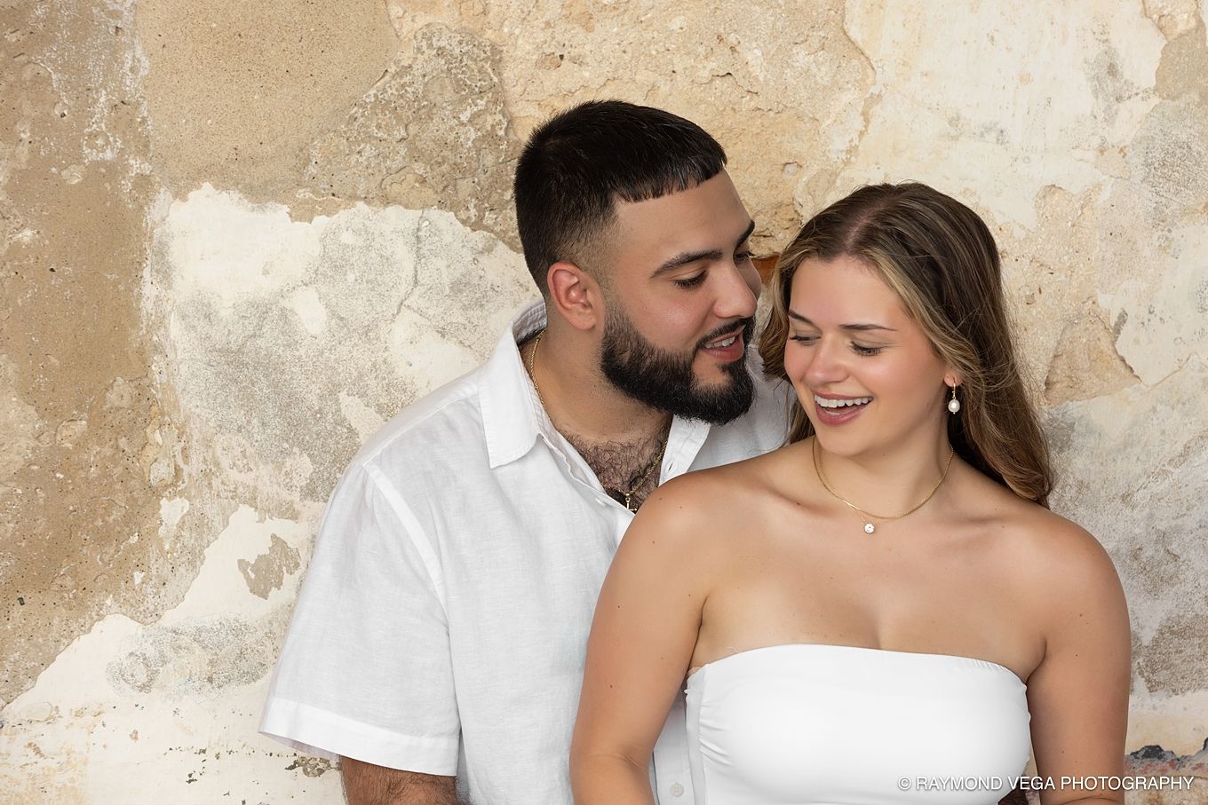 Romantic couple in white outfits sharing a tender moment against a rustic stone wall outdoors.