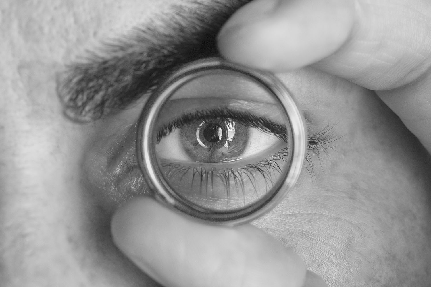 Black and white close-up of a human eye viewed through a magnifying glass held by fingers. Raymond Vega Photography & Cinematography
