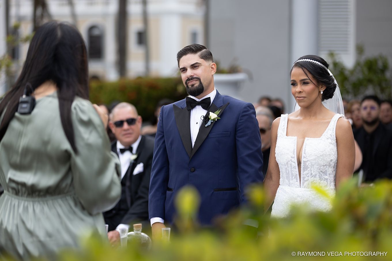 Bride and groom stand at outdoor wedding ceremony, groom in black tuxedo with yellow boutonniere, bride in white lace gown. Antiguo Casino