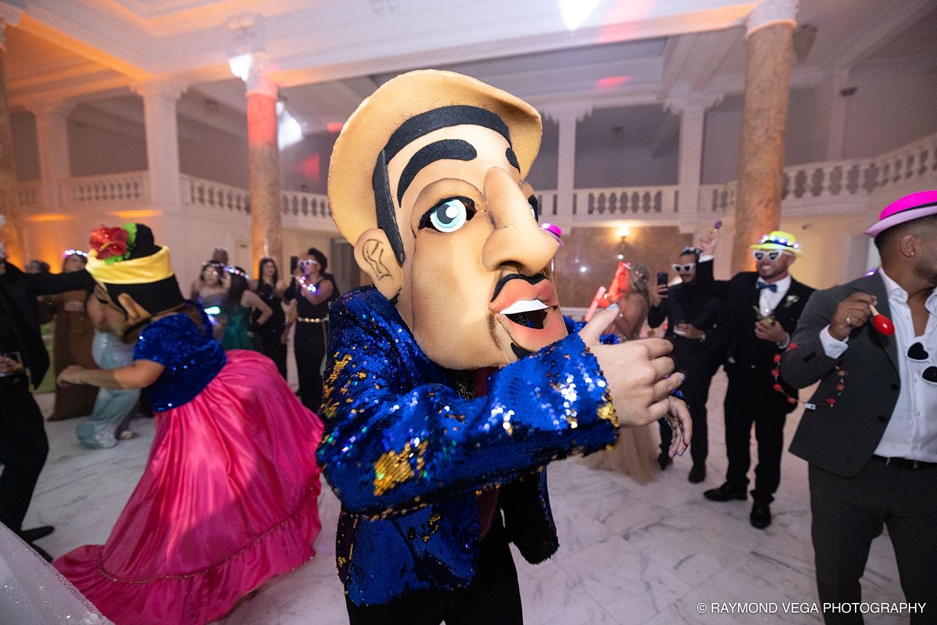 Person in oversized caricature head costume in blue sequin jacket dancing at a colorful masquerade party event. PLENEROS PUERTO RICO