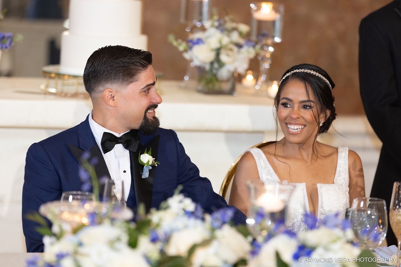 Bride and groom smiling and laughing together at wedding reception sweetheart table with blue and white floral centerpieces.