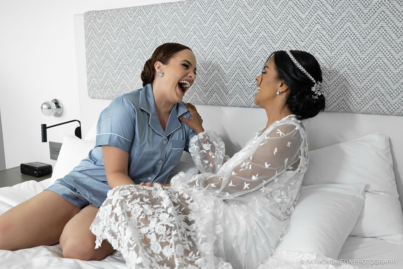 Two women laughing joyfully on a white bed, one in a blue pajama set and the other in a floral bridal robe. Hotel la Concha Wedding