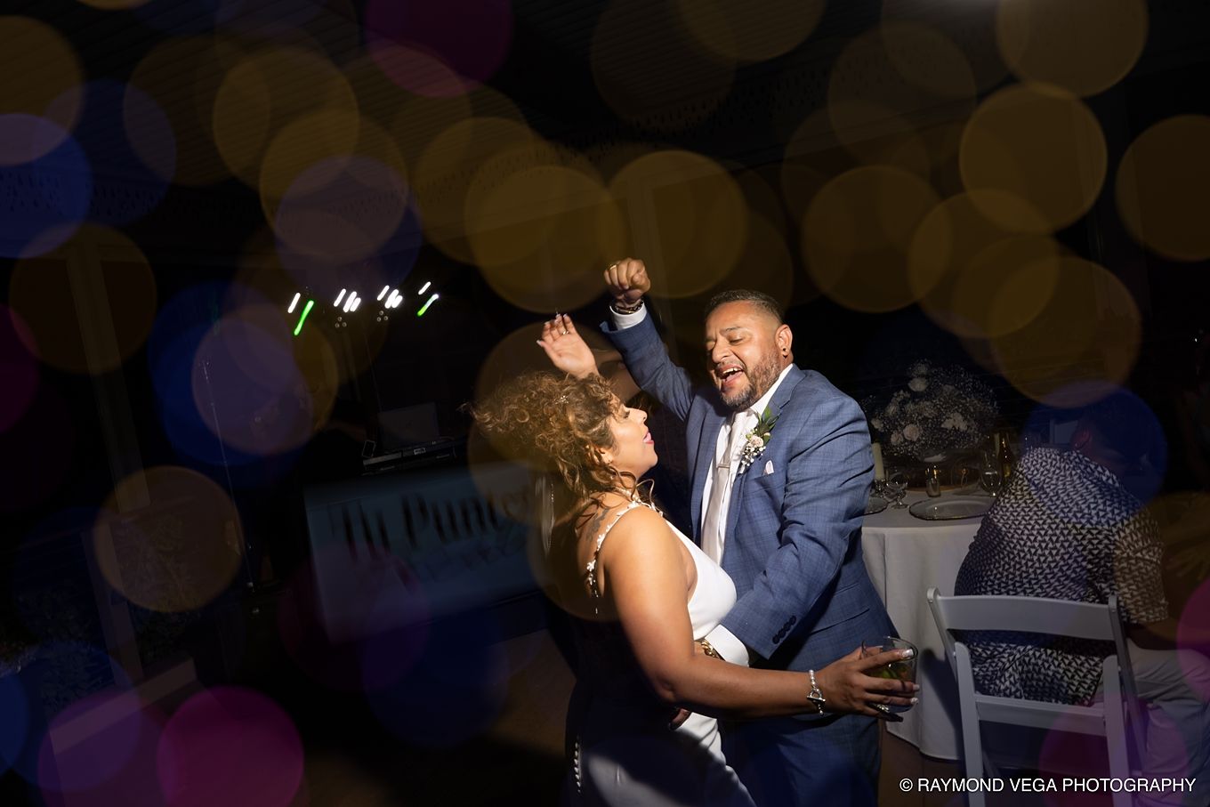 Joyful couple dancing at wedding reception with colorful bokeh lights in background.
