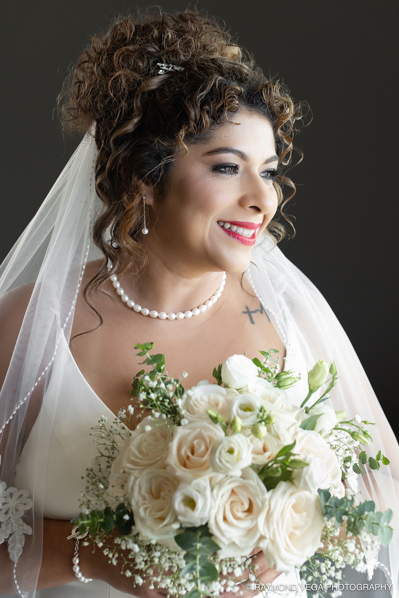 Smiling bride with curly hair, pearl necklace, cross tattoo, white veil, and bouquet of white roses on dark background.