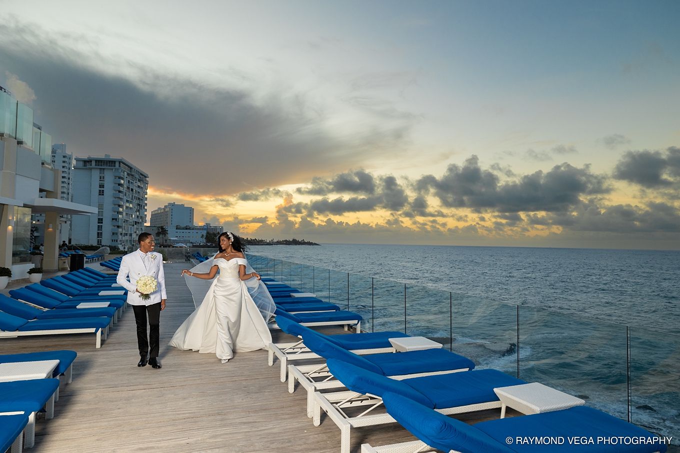 Bride and groom walk along oceanfront deck at sunset, blue lounge chairs lining the path at a Cancun resort wedding.