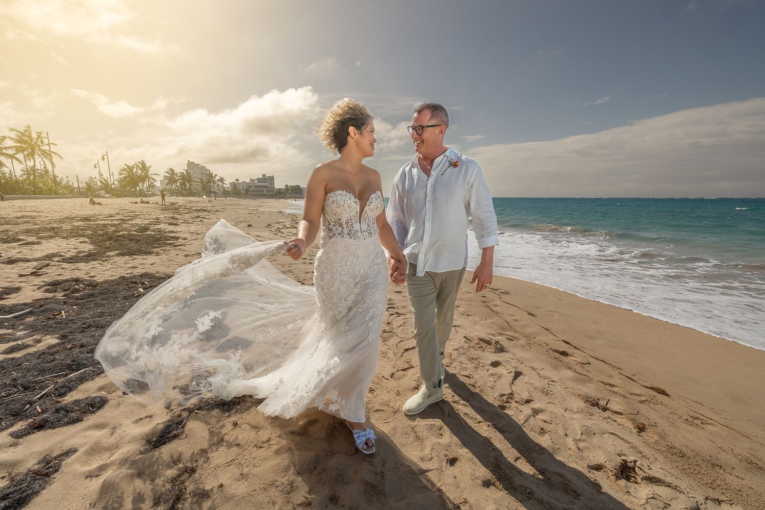Bride and groom walking hand in hand on a sunny beach, wedding veil flowing in the breeze with ocean views.