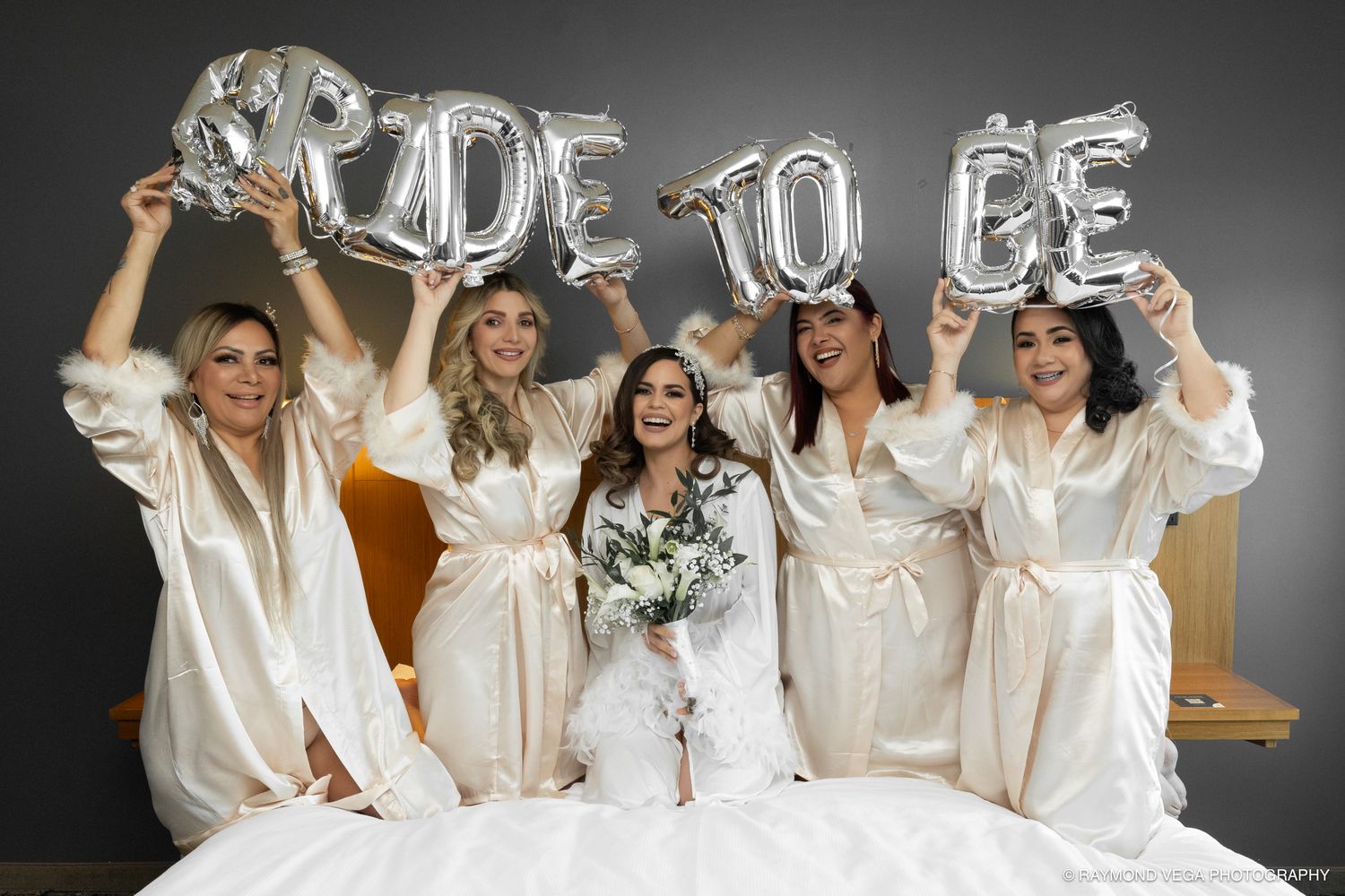 Bride in wedding dress with bridesmaids in satin robes holding silver BRIDE TO BE balloons on a bed.