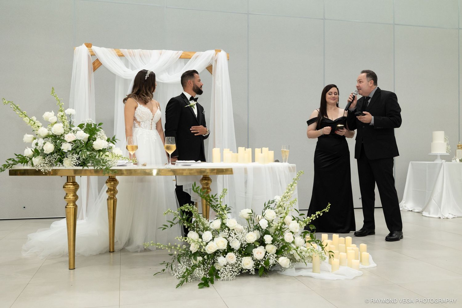 Elegant wedding ceremony with couple at gold altar, white floral arrangements, candles, and guests in a bright venue.