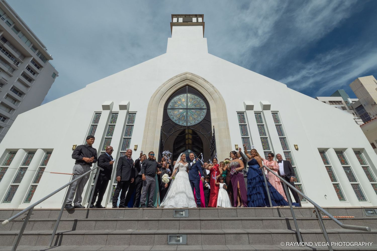 Wedding party posing on steps outside a white church with gothic arch doorway under blue sky.