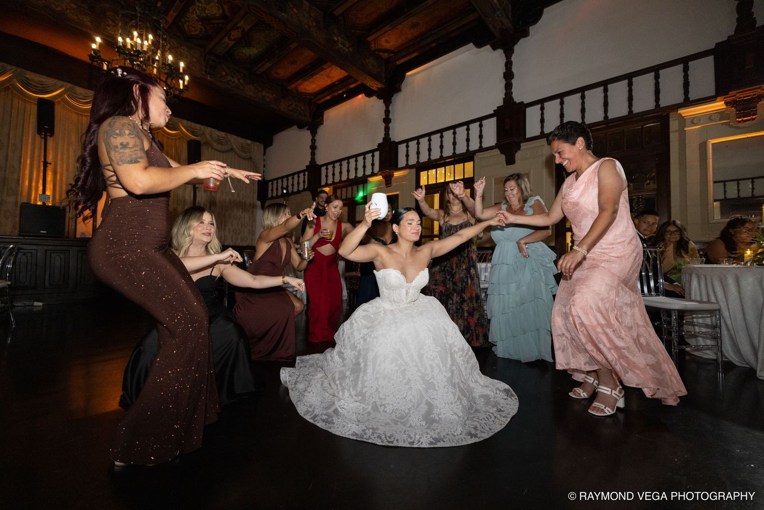 Bride in white gown dances joyfully with bridesmaids in colorful dresses at elegant wedding reception.
