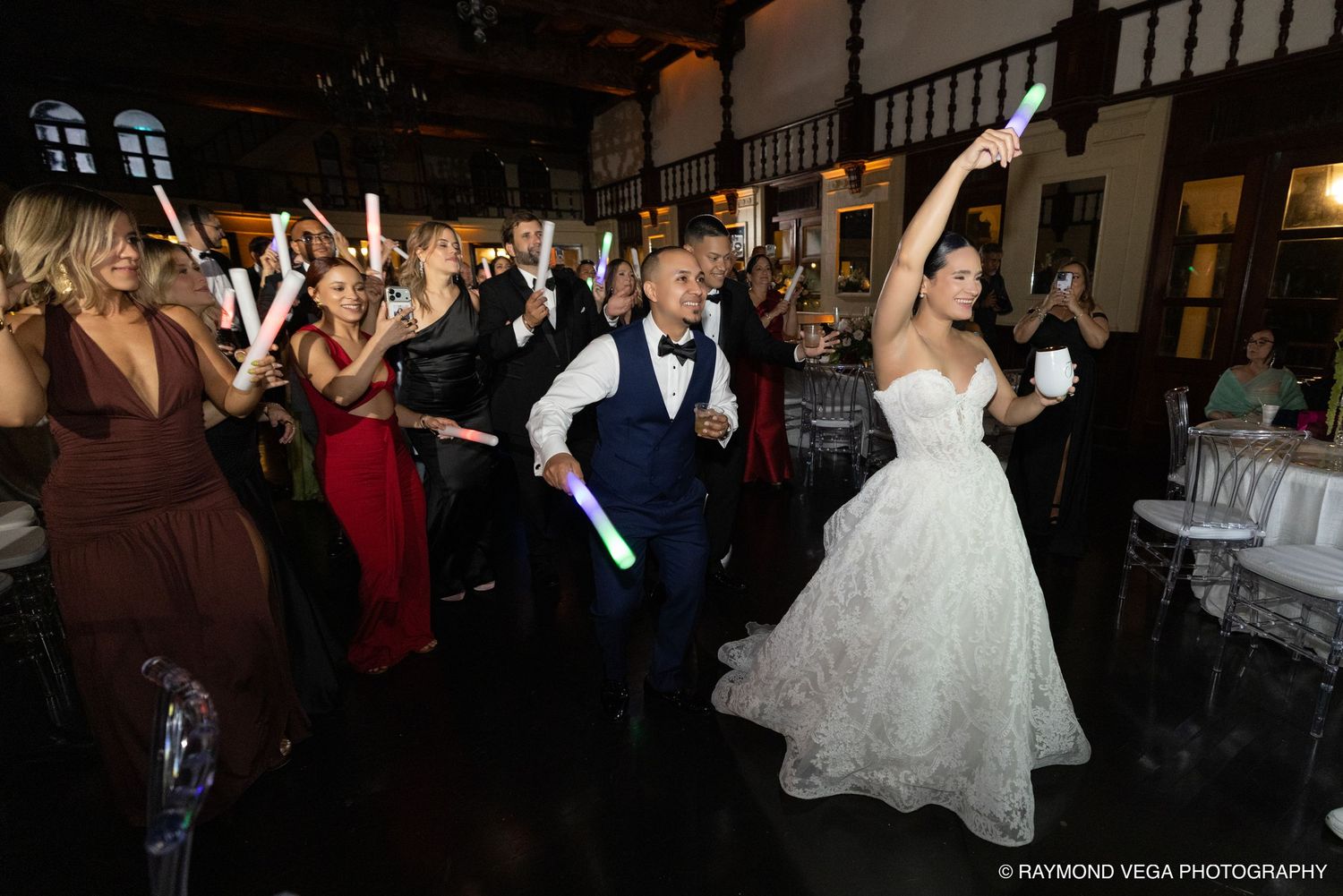 Bride and groom dancing joyfully with guests holding glow sticks at a lively indoor wedding reception celebration.
