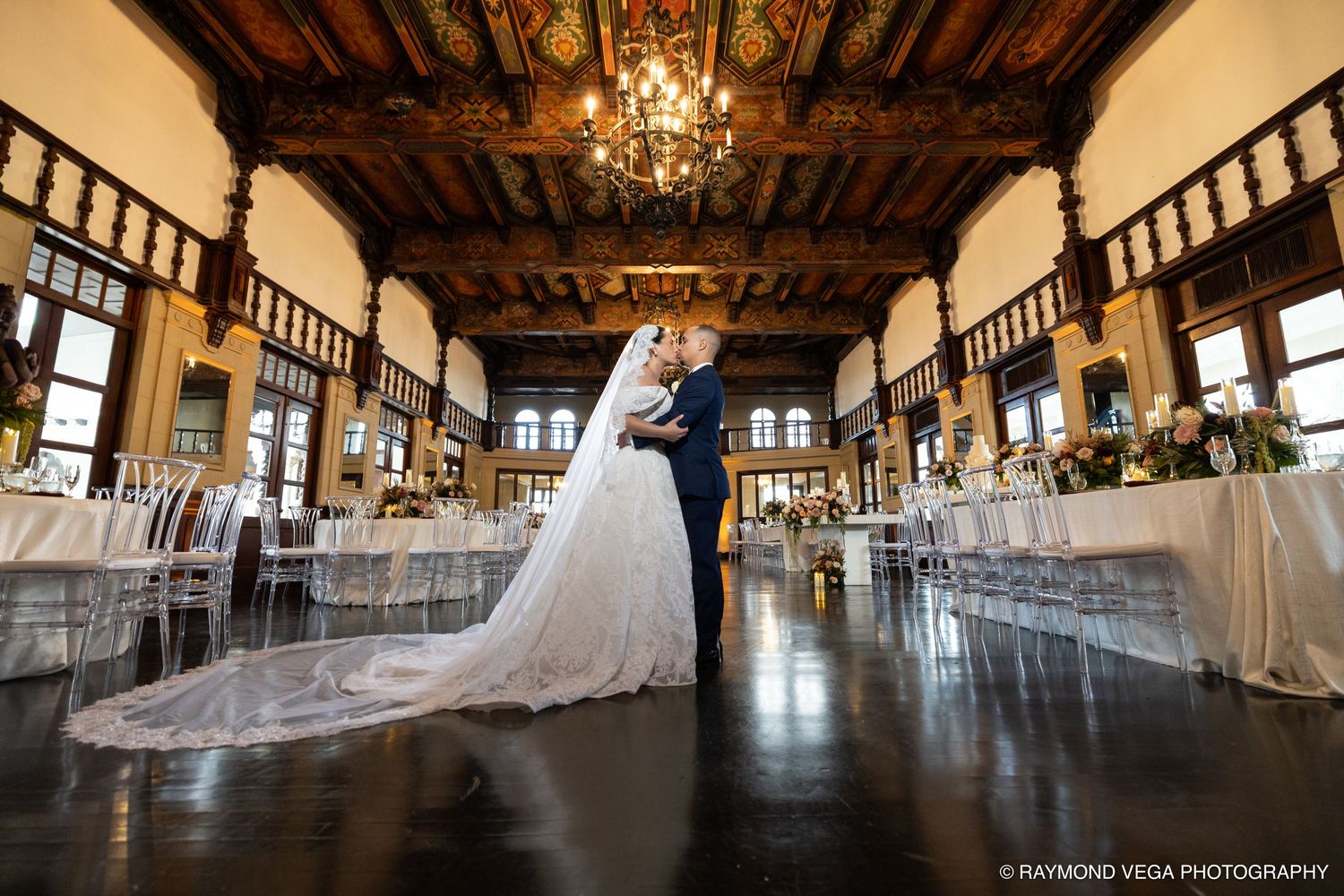 Bride and groom share romantic embrace in elegant ballroom with chandelier and reception tables at wedding venue.