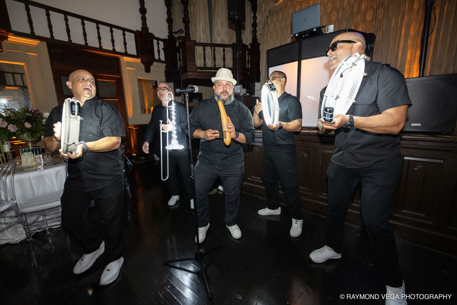 Five musicians in black attire perform with brass instruments at an elegant indoor event venue.